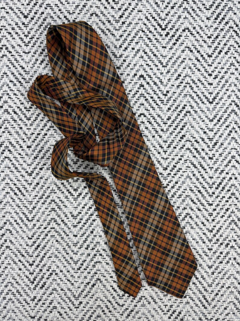 Puede incluir: Una corbata con un patr&oacute;n de cuadros en tonos naranja quemado, negro y beige. La tela tiene una textura suave y el dise&ntilde;o consiste en l&iacute;neas diagonales que se cruzan formando un motivo de cuadr&iacute;cula cl&aacute;sico a lo largo de todo el accesorio.