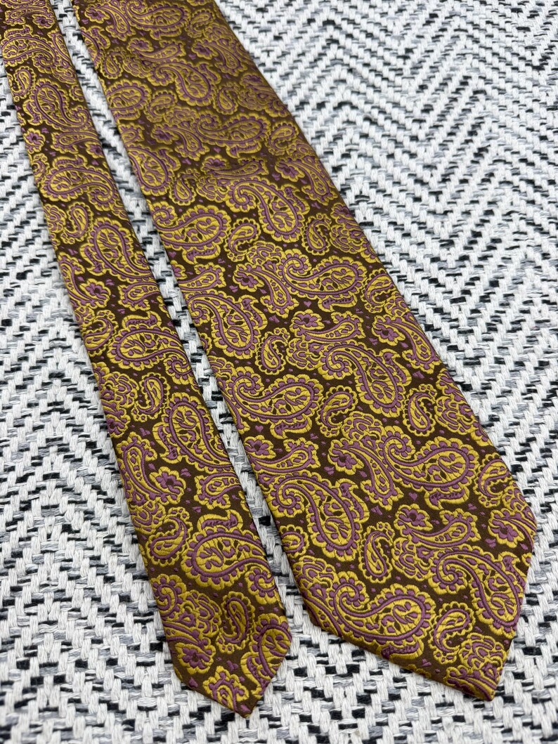 May include: A necktie featuring a detailed paisley pattern in shades of brown, gold, and purple. The fabric displays a repeating intricate design of teardrop-shaped motifs across the entire surface of the tie, including the narrow and wide ends.