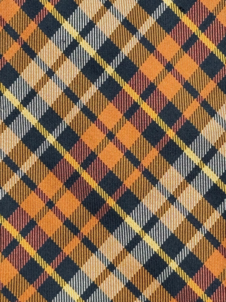 Puede incluir: Una tela con un patr&oacute;n de cuadros diagonal en tonos negro, naranja, amarillo y blanco. El dise&ntilde;o consiste en l&iacute;neas y bloques de color que se cruzan para crear una cuadr&iacute;cula geom&eacute;trica repetitiva en toda la superficie del material.