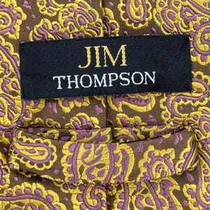 May include: A brown necktie featuring a repeating paisley pattern in yellow and purple. A black fabric label is stitched onto the center of the tie, displaying the text JIM THOMPSON in gold lettering. The tie includes a fabric loop on the back.