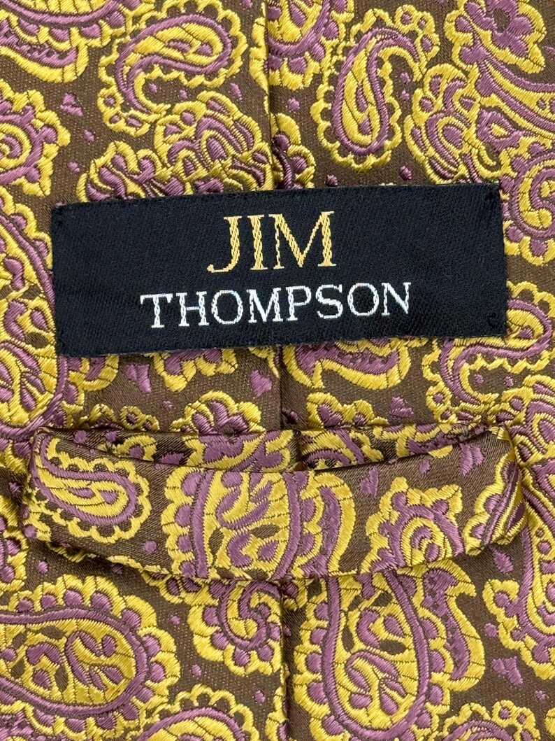 May include: A brown necktie featuring a repeating paisley pattern in yellow and purple. A black fabric label is stitched onto the center of the tie, displaying the text JIM THOMPSON in gold lettering. The tie includes a fabric loop on the back.