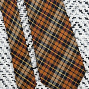 Puede incluir: Una corbata con un patr&oacute;n de cuadros con l&iacute;neas que se cruzan en tonos naranja quemado, negro y rayas blancas finas. La tela muestra una textura tejida consistente en todo el dise&ntilde;o, destacando la orientaci&oacute;n diagonal del tart&aacute;n.