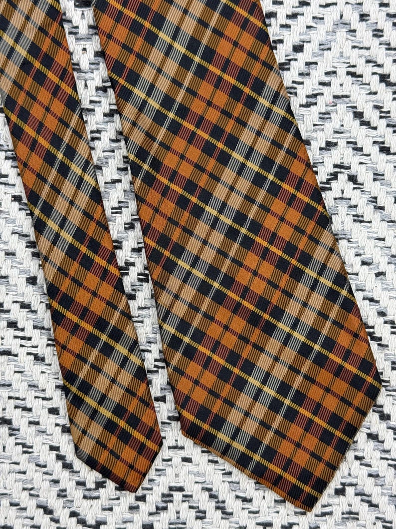Puede incluir: Una corbata con un patr&oacute;n de cuadros con l&iacute;neas que se cruzan en tonos naranja quemado, negro y rayas blancas finas. La tela muestra una textura tejida consistente en todo el dise&ntilde;o, destacando la orientaci&oacute;n diagonal del tart&aacute;n.