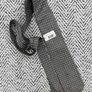 May include: A black and white checkered tie with a small tag attached. The tie is made of a woven fabric and is lying on a textured, patterned surface. The tie is a classic accessory.