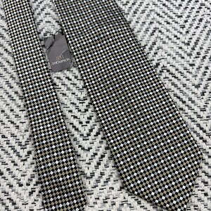 May include: A black, white, and tan checkered necktie with a tag that reads "Jim Thompson". The tie is lying on a textured, black and white chevron patterned surface. The tie is a classic accessory.