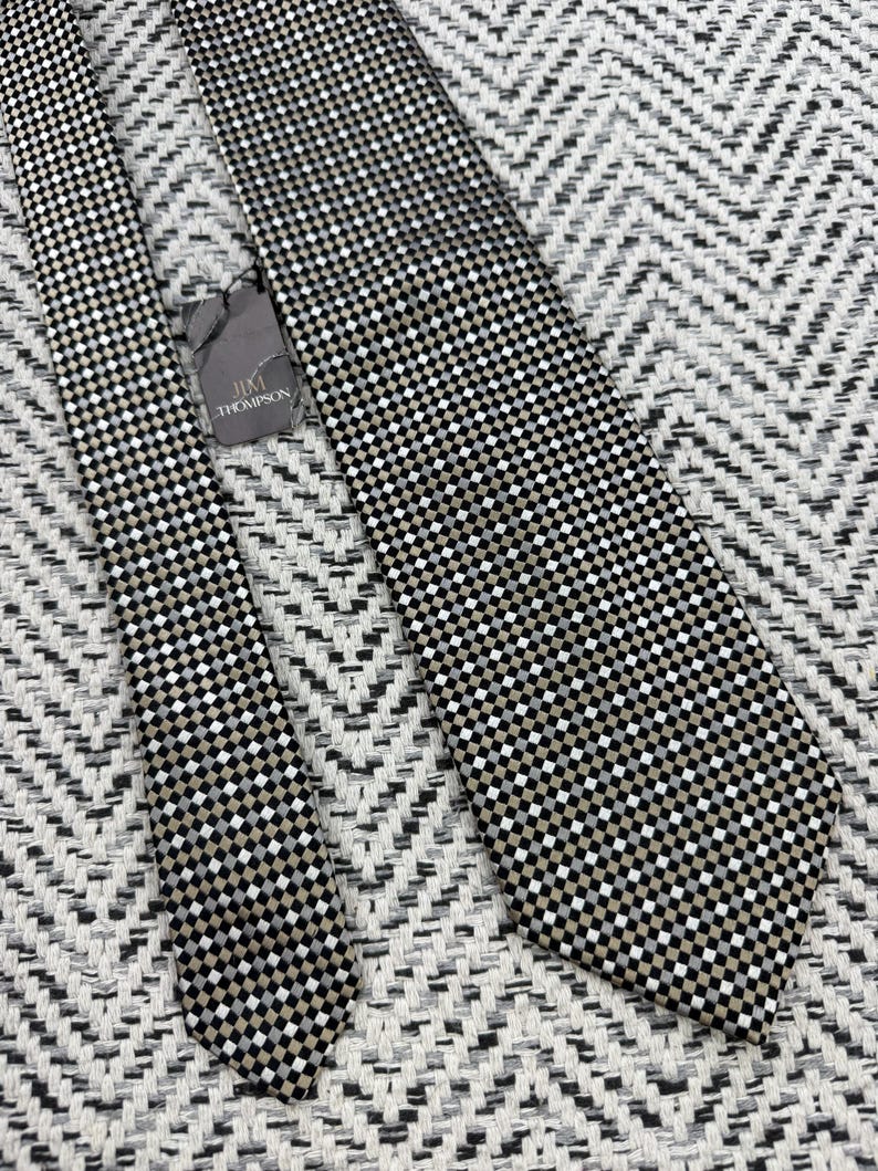 May include: A black, white, and tan checkered necktie with a tag that reads "Jim Thompson". The tie is lying on a textured, black and white chevron patterned surface. The tie is a classic accessory.