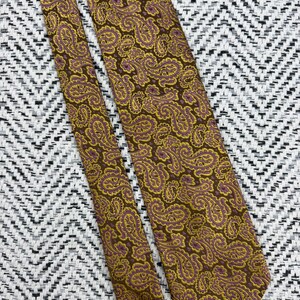 May include: A necktie featuring a brown background with an intricate paisley pattern in shades of mustard yellow and muted purple. The fabric has a woven texture, and the design covers the entire length of the tie, including the pointed ends.
