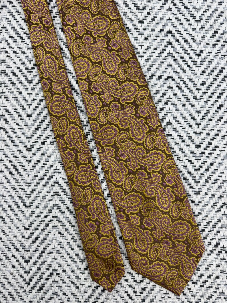May include: A necktie featuring a brown background with an intricate paisley pattern in shades of mustard yellow and muted purple. The fabric has a woven texture, and the design covers the entire length of the tie, including the pointed ends.
