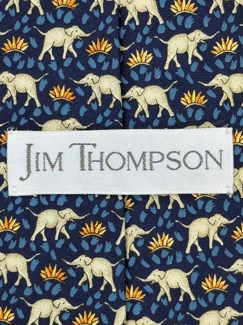 May include: A navy blue tie featuring a repeating pattern of beige elephants, orange and blue accents. The label reads "JIM THOMPSON".