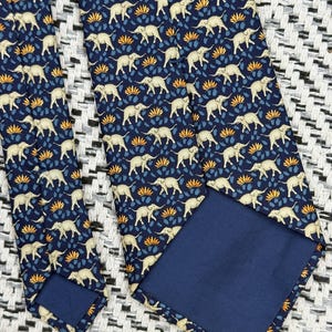 May include: A navy blue tie with a repeating pattern of cream elephants, blue leaves, and orange floral details. The tie has a solid navy blue backing and is displayed on a textured black and white surface.