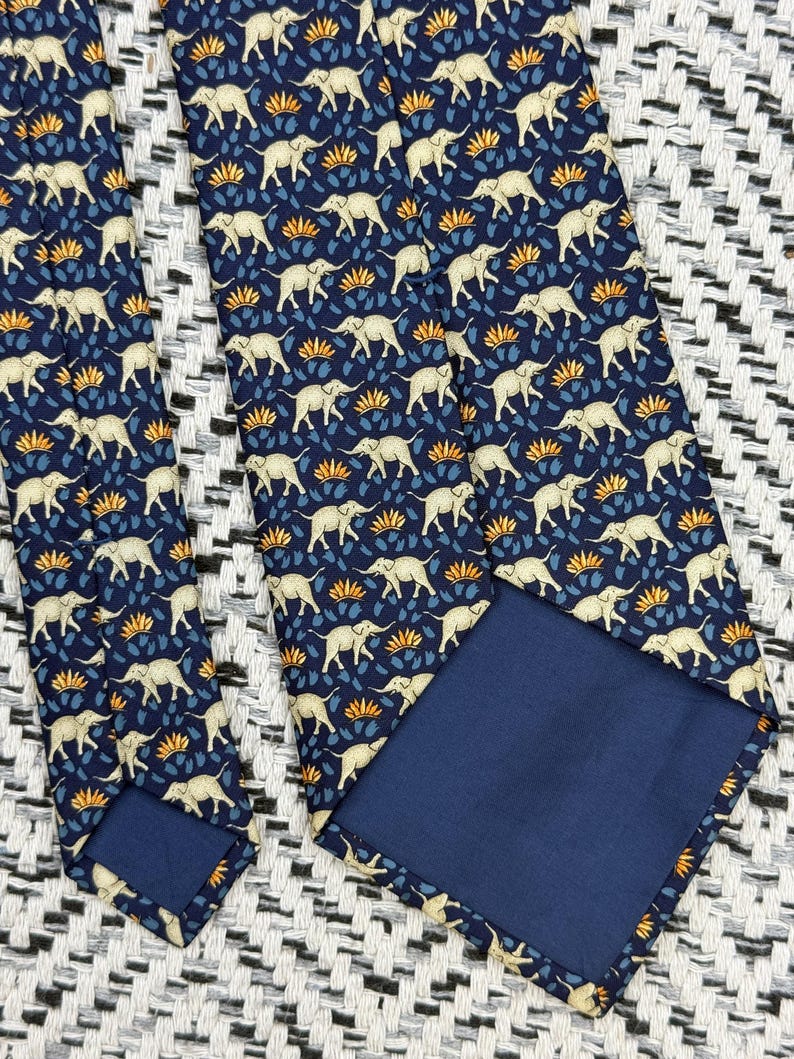 May include: A navy blue tie with a repeating pattern of cream elephants, blue leaves, and orange floral details. The tie has a solid navy blue backing and is displayed on a textured black and white surface.