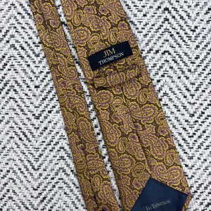 May include: A necktie featuring a brown, yellow, and purple paisley pattern. The tie includes a black label with the text JIM THOMPSON in gold lettering and a dark blue fabric lining at the tip with the text JIM THOMPSON printed in white.