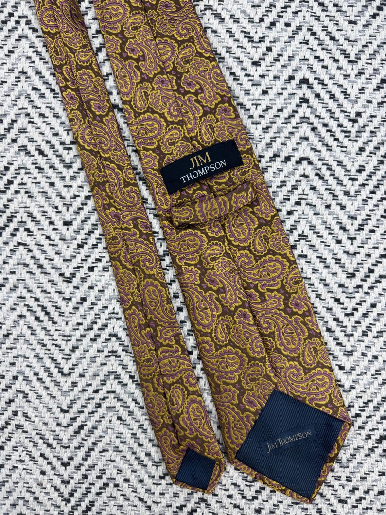 May include: A necktie featuring a brown, yellow, and purple paisley pattern. The tie includes a black label with the text JIM THOMPSON in gold lettering and a dark blue fabric lining at the tip with the text JIM THOMPSON printed in white.