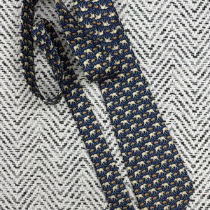 May include: A navy blue tie featuring a repeating pattern of cream elephants and orange floral details. The tie is made of a smooth fabric and is displayed on a textured black and white background.