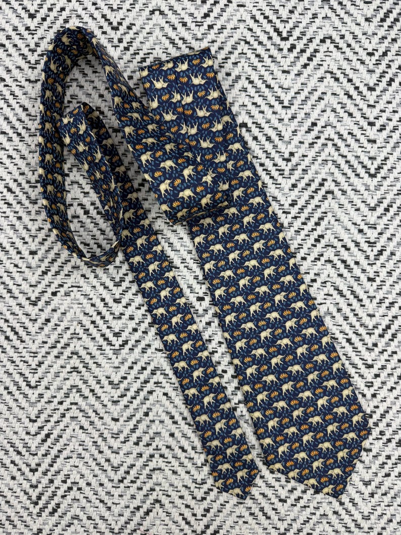 May include: A navy blue tie featuring a repeating pattern of cream elephants and orange floral details. The tie is made of a smooth fabric and is displayed on a textured black and white background.