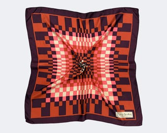 May include: A square silk scarf featuring a geometric design in red, orange, and cream hues. It has a dark purple border and a label in the bottom right corner that reads "Pierre Cardin".
