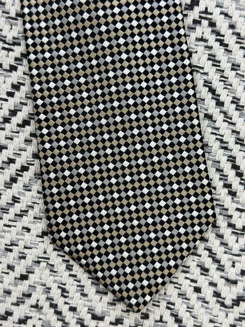May include: A patterned necktie with a repeating checkered design in black, white, and tan. The tie is made of a smooth fabric and is displayed on a textured surface.