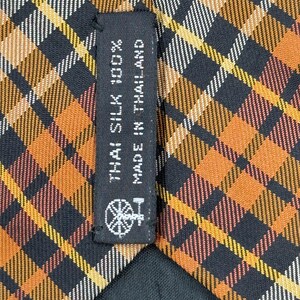Puede incluir: Una corbata con un patr&oacute;n de cuadros en tonos naranja, negro, blanco y amarillo. Una etiqueta de tela negra est&aacute; cosida en la parte posterior, mostrando el texto THAI SILK 100% MADE IN THAILAND y un peque&ntilde;o icono gr&aacute;fico de una rueca.