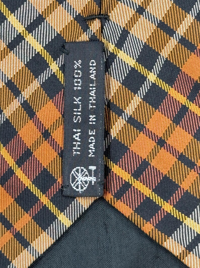 Puede incluir: Una corbata con un patr&oacute;n de cuadros en tonos naranja, negro, blanco y amarillo. Una etiqueta de tela negra est&aacute; cosida en la parte posterior, mostrando el texto THAI SILK 100% MADE IN THAILAND y un peque&ntilde;o icono gr&aacute;fico de una rueca.