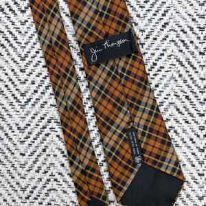 Puede incluir: Una corbata con un patr&oacute;n de cuadros en tonos naranja, negro y blanco. La corbata incluye una etiqueta negra con el texto Jim Thompson y una etiqueta negra m&aacute;s peque&ntilde;a con el texto Thai Silk 100% Made in Thailand, en la parte posterior.