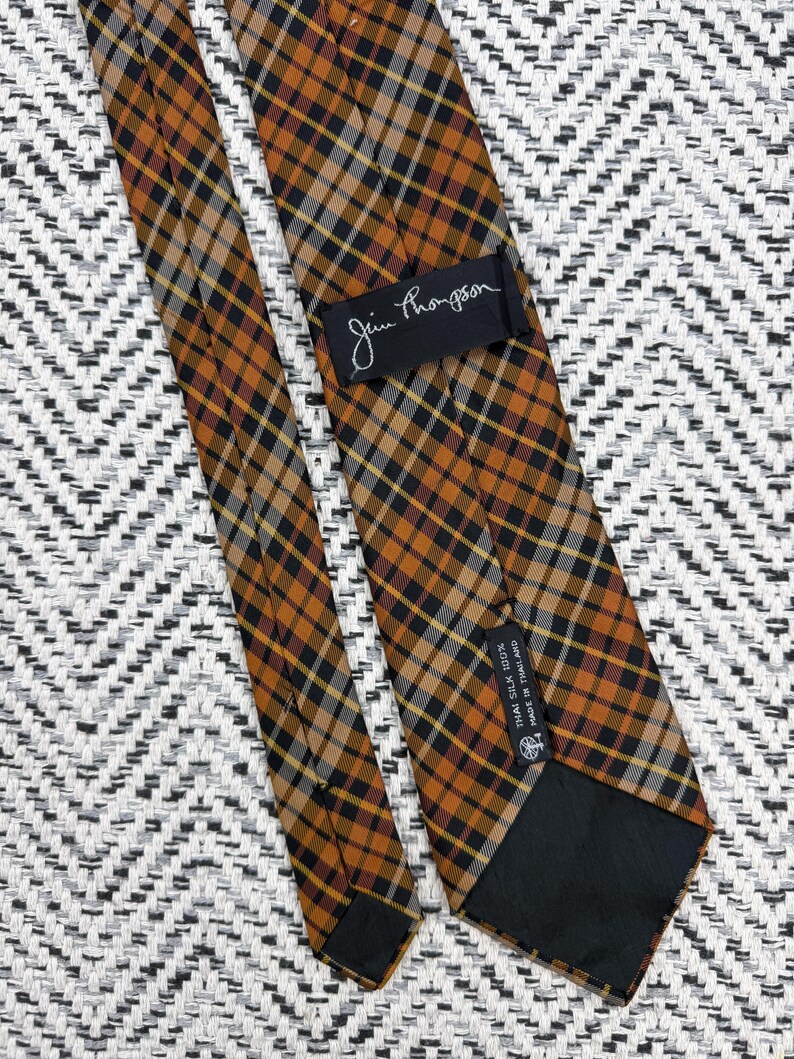 Puede incluir: Una corbata con un patr&oacute;n de cuadros en tonos naranja, negro y blanco. La corbata incluye una etiqueta negra con el texto Jim Thompson y una etiqueta negra m&aacute;s peque&ntilde;a con el texto Thai Silk 100% Made in Thailand, en la parte posterior.