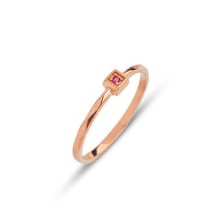 Può includere: Un delicato anello in oro rosa con una piccola pietra preziosa di rubino di forma quadrata incastonata al centro.