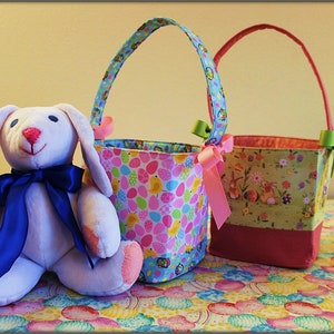 May include: Two Easter baskets with fabric handles and colorful patterns. One basket has a pink and blue pattern with eggs and birds. The other basket has a green and pink pattern with bunnies and flowers. A white plush bunny with blue ribbon sits in front of the baskets.