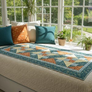 Op de afbeelding: Een dagbed met een crèmekleurige doorgestikte sprei, bedekt met een rechthoekige quiltloper in geometrische patronen van petrol, oranje en crème. Vier vierkante sierkussens in effen petrol en oranje bloemenprint zijn langs de achterkant geplaatst.