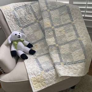 May include: A white and gray quilted baby blanket with a yellow floral pattern. A white stuffed lamb is sitting on a beige armchair.