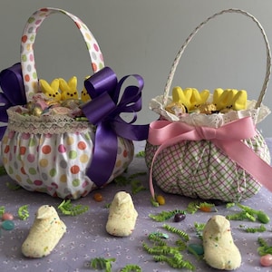 May include: Two Easter baskets filled with yellow candy peeps and wrapped in fabric. The larger basket is white with colorful polka dots and a purple bow. The smaller basket is green and white plaid with a pink bow.