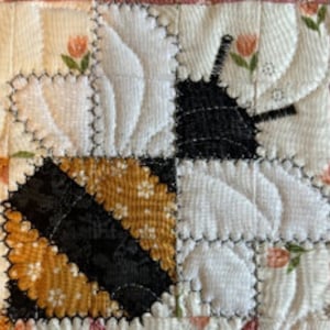 May include: A close-up of a bee quilt block made from fabric scraps. The bee is yellow, black, and white with a black and white striped body. The bee is surrounded by white fabric with a floral pattern.