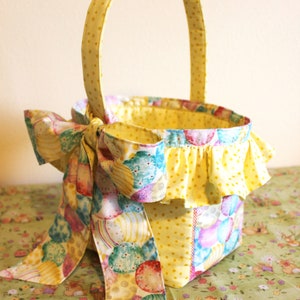 May include: A yellow fabric Easter basket with a ruffled edge and a bow made from a colorful fabric with a pattern of Easter eggs. The basket is sitting on a green and white floral tablecloth.