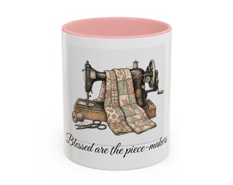 May include: White ceramic mug with a pink interior. The mug features an illustration of a vintage sewing machine with a patchwork quilt draped over it. The text "Blessed are the piece-makers" is printed on the mug.