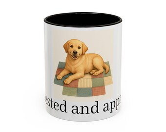 Könnte beinhalten: Weißer Keramikbecher mit schwarzem Innenrand. Der Becher zeigt einen goldenen Labrador-Welpen, der auf einer Patchwork-Steppdecke liegt. Der Text "tested and app" ist auf dem Becher aufgedruckt.