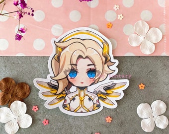 May include: A sticker featuring a cartoon illustration of a female character with blonde hair, blue eyes, and a halo. She is wearing a white and gold suit with wings. The sticker is on a pink and white polka dot background.