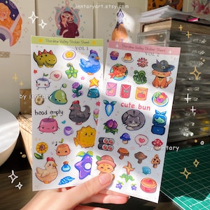 May include: Two sticker sheets featuring illustrations from the video game Stardew Valley. The first sheet is labeled "Vol. 1" and features characters and items from the game, including a blue bird, a green slime, a chicken, and a sunflower. The second sheet is labeled "Vol. 2" and features characters and items from the game, including a cat wearing a witch's hat, a mushroom, and a sushi roll.