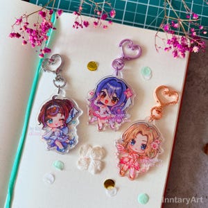 May include: Three clear acrylic keychains with anime-style characters. Each keychain has a heart-shaped clasp. The characters have large eyes and colourful hair, and are dressed in detailed outfits with floral accents. The text "by inntary do not use" is printed on each keychain.