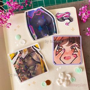 May include: Three holographic stickers featuring characters from the video game Overwatch. The stickers are on a white background with a light blue and white floral pattern. The text "by inntary" appears on two of the stickers.