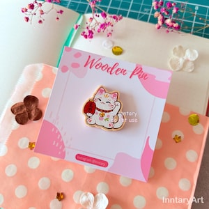May include: A wooden pin with a Maneki-neko cat design. The cat is white with pink details, holding a red coin bag. The pin is on a pink card with the text "Wooden Pin" and "Instagram @Inntary".