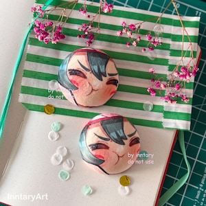May include: Two round, plush character buttons with a smiling face design, featuring teal hair and a red headband. The buttons are on a notebook with green and white striped paper and pink flowers. The text "by inntary do not use" is visible.