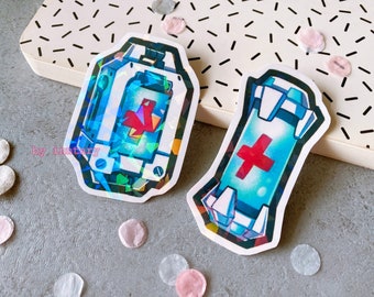 May include: Two holographic stickers featuring a medical kit and a medical syringe. Both stickers have a blue and white design with a red cross.