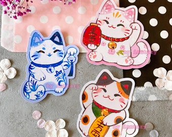 Op de afbeelding: Drie schattige cartoon gelukkatten stickers met glitter accenten. De katten staan in verschillende poses, met een gouden munt, een rode geldbeurs en een bel. De stickers zijn op een roze en witte stippen achtergrond.