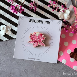 Puede incluir: Un pin de madera con un diseño de flor de loto rosa. El pin está sobre una tarjeta blanca con el texto "WOODEN PIN" y el nombre de Instagram "@Inntary_". El fondo incluye flores decorativas y papel estampado.