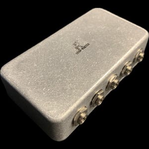 Puede incluir: Una caja de metal plateada con el texto "LOOP-MASTER" impreso en la parte superior. La caja tiene cinco conectores metálicos plateados en el lateral.