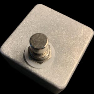 May include: A silver-colored, square-shaped effects pedal with a single, round, silver-colored footswitch. The footswitch is surrounded by a white washer. The pedal has a textured surface and is set against a black background.