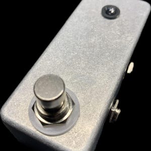 May include: A silver-colored rectangular guitar effects pedal with a single footswitch and a small black screw on top. The pedal has a textured surface and is designed for use with electric guitars.
