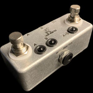 Peut inclure: Pédale d'effets de guitare Loop Master argentée avec deux pédales et des prises d'entrée/sortie. La pédale a des boutons noirs et des accents blancs, avec du texte indiquant les entrées et les sorties.