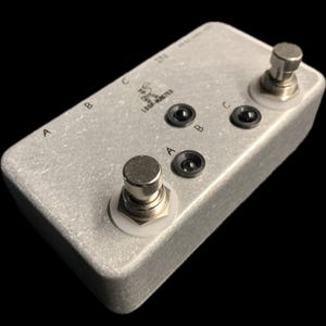 Peut inclure: Pédale d'effets de guitare Loop Master argentée avec trois interrupteurs au pied étiquetés A, B et C, et deux prises d'entrée/sortie. La pédale a une surface texturée et une finition argentée. Le texte "Loop Master" est imprimé sur le dessus.