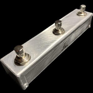May include: A brushed aluminum guitar pedal with three silver footswitches. The rectangular enclosure has a textured surface and the text "Loop Master" is visible. The pedal is designed for use with electric guitars.