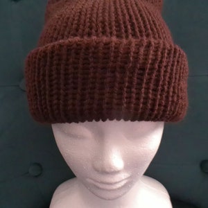 Puede incluir: Un gorro de punto marr&oacute;n sobre una cabeza de maniqu&iacute; blanca. El gorro est&aacute; hecho de un hilo grueso y texturizado y tiene un dise&ntilde;o simple y cl&aacute;sico.